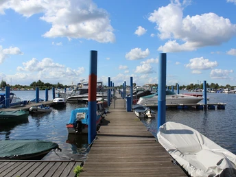 Holzsteg führt durch eine Boots-Marina mit verschiedenen Motorbooten bei blauem Himmel und Wolken.