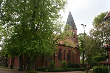 St. Vitus-Kirche Schinna, eine Backsteinkirche mit hohem Turm, umgeben von Bäumen, Himmel bedeckt.