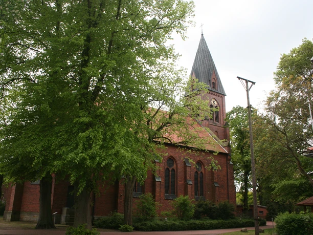 St. Vitus-Kirche Schinna, eine Backsteinkirche mit hohem Turm, umgeben von Bäumen, Himmel bedeckt.
