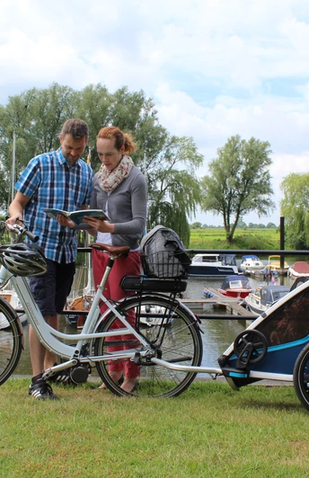 Ein Paar mit Fahrradanhänger betrachtet eine Karte am grünen Ufer des Achimer Yachthafens mit Booten.