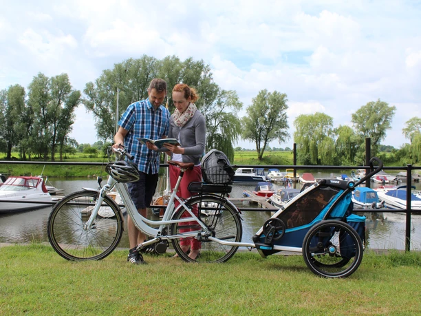 A couple with a bicycle trailer look at a map on the green shore of the Achim marina with boats.