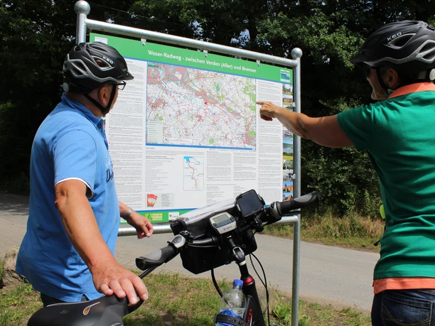 Cyclists look at an information board on the Weser cycle path under trees in sunny weather.