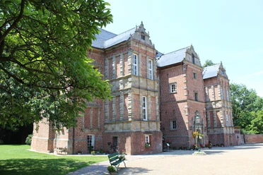 Schloss Erbhof Historisches Backsteinschloss mit grüner Umgebung und blauen Türen, beleuchtet von sommerlichem Sonnenlicht.