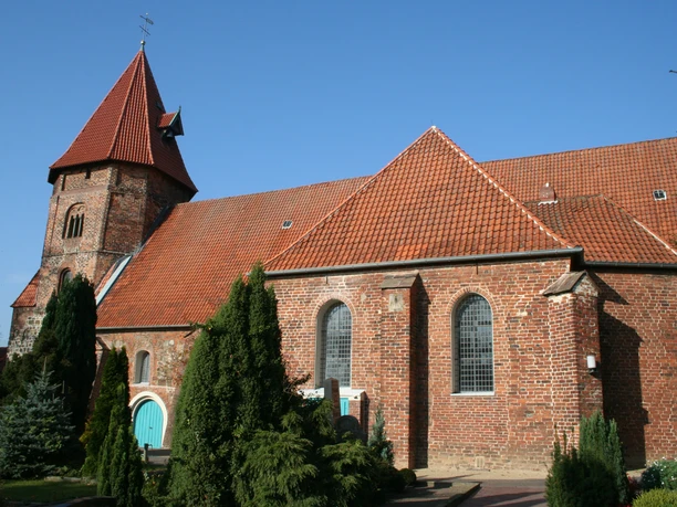 Backsteinkirche mit rotem Satteldach, spitzem Turm und grüner Holztür, umgeben von Bäumen und Pflanzen.