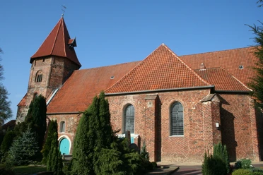 St. Laurentius-Kirche Backsteinkirche mit rotem Satteldach, spitzem Turm und grüner Holztür, umgeben von Bäumen und Pflanzen.