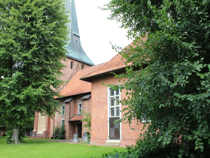 Backsteinkirche mit grünem Spitzdach, flankiert von großen Laubbäumen, auf einer gepflegten Rasenfläche.