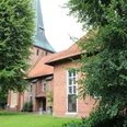 St.-Marcellus-Kirche Asendorf Backsteinkirche mit grünem Spitzdach, flankiert von großen Laubbäumen, auf einer gepflegten Rasenfläche.