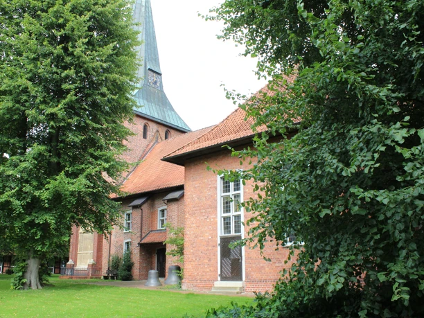 St.-Marcellus-Kirche Asendorf Backsteinkirche mit grünem Spitzdach, flankiert von großen Laubbäumen, auf einer gepflegten Rasenfläche.