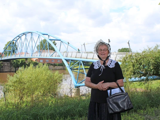 Eine Frau in historischer Tracht steht vor einer blauen Brücke an einem Flussufer in Nienburg.