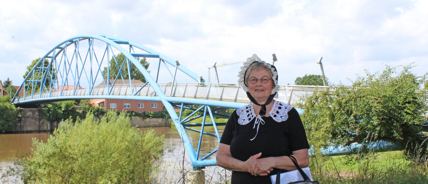 Gewandete Stadtführerin Nienburg Eine Frau in historischer Tracht steht vor einer blauen Brücke an einem Flussufer in Nienburg.