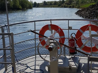 An Bord Ein Boot auf einem ruhigen Fluss mit Rettungsringen und grün bewachsenen Ufern im Hintergrund.