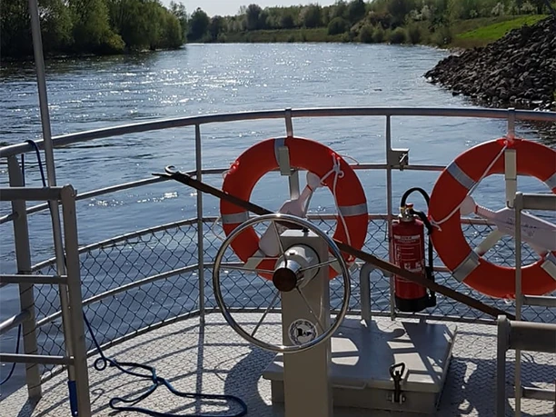 An Bord Ein Boot auf einem ruhigen Fluss mit Rettungsringen und grün bewachsenen Ufern im Hintergrund.