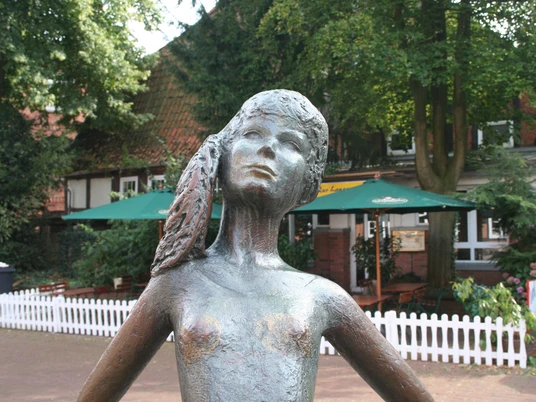 Bronzestatue eines Mädchens mit Zöpfen im Freien, umgeben von Bäumen und einem Café im Hintergrund.