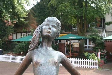 Kleine Nienburgerin Bronzestatue eines Mädchens mit Zöpfen im Freien, umgeben von Bäumen und einem Café im Hintergrund.