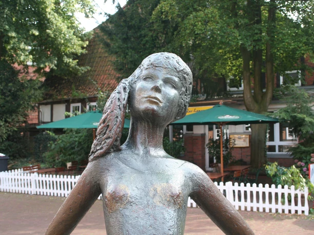 Kleine Nienburgerin Bronzestatue eines Mädchens mit Zöpfen im Freien, umgeben von Bäumen und einem Café im Hintergrund.
