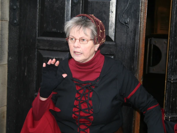 Ältere Frau in mittelalterlicher Kleidung steht vor einer Holztür und hält die Hand erhoben.
