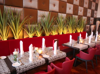 (Foto Frank Wilde) Auf dem Bild ist eine lange rote Sitzbank zu sehen, an der mehrere Tische mit roten Stühlen angeordnet sind. Alle Tische sind eingedeckt. Vor der Wand stehen dekorative Sträucher.