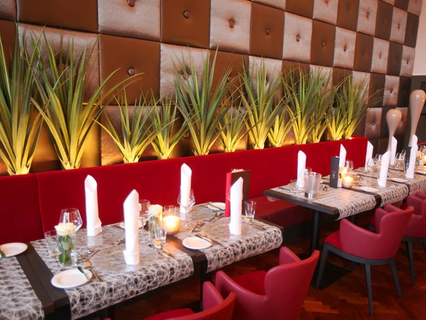 (Foto Frank Wilde) Auf dem Bild ist eine lange rote Sitzbank zu sehen, an der mehrere Tische mit roten Stühlen angeordnet sind. Alle Tische sind eingedeckt. Vor der Wand stehen dekorative Sträucher.