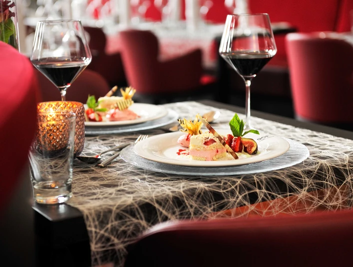 Palmengarten Desert (c) GOP Variete.jpg Auf dem Bild ist ein gedeckter Tisch zu sehen, der mit zwei Tellern, zwei Gläsern Rotwein und einem Kerzenlicht dekoriert ist. Im Hintergrund sieht man Stühle mit einem roten Stoff.