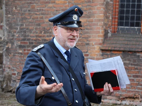 Ein Gästeführer in historischer Polizeiuniform spricht gestikulierend vor einer Backsteinwand.