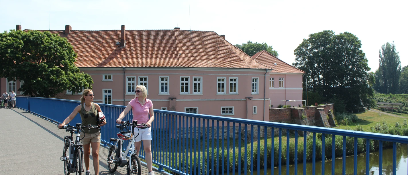 Radfahrer am Grafenschloss Hoya Zwei Radfahrer auf einer Brücke vor dem historischen Grafenschloss Hoya an einem sonnigen Tag.