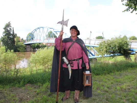 Ein Mann in traditioneller Nachtwächterkleidung steht vor einer Brücke über einen Fluss.