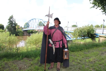 Nienburger Nachtwächter Ein Mann in traditioneller Nachtwächterkleidung steht vor einer Brücke über einen Fluss.