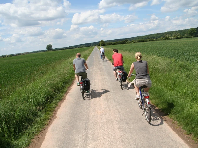 Radfahrer auf einem schmalen Weg entlang grüner Felder, blauer Himmel mit weißen Wolken oben.