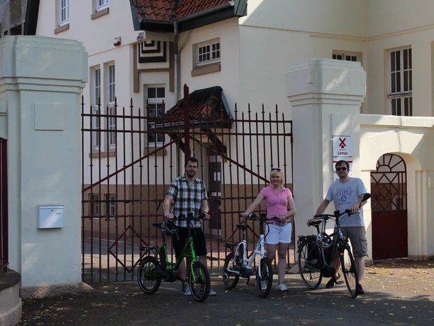 Three people with bicycles stand in front of the historic gates of the Eystrup mustard factory in the sunshine.