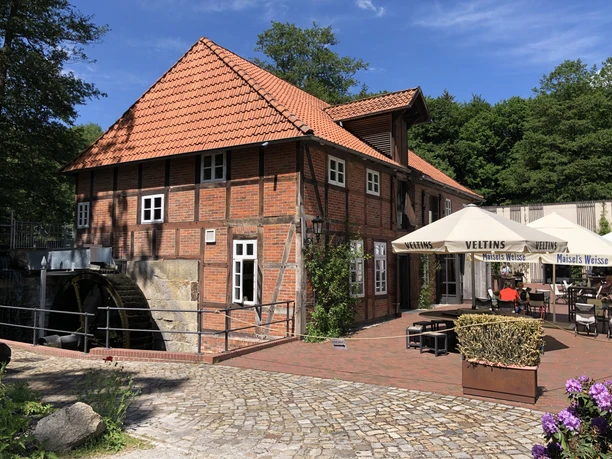 Historic mill with red half-timbering, surrounded by trees and parasols in the outdoor dining area.