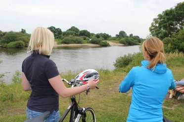 Two women stand on the banks of the Weser with bicycles, looking out over the calm river and nature.