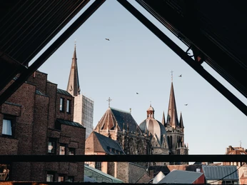 Der Aachener Dom ragt in den Himmel, eingefasst in ein Gerüst aus alten Gebäuden und modernen Strukturen. Seine gotischen Türme thronen über der Umgebung und fangen das Licht des klaren Himmels.