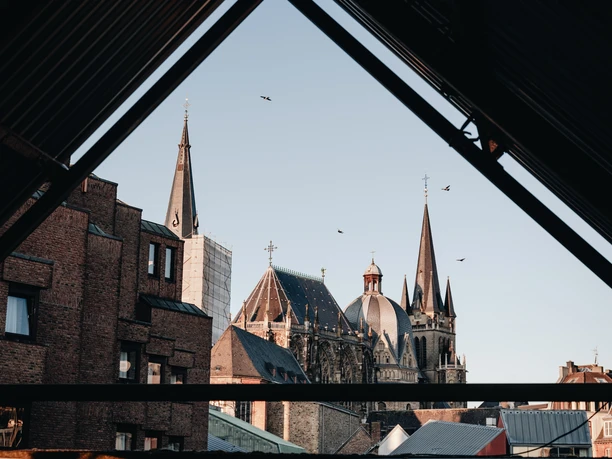 Der Aachener Dom ragt in den Himmel, eingefasst in ein Gerüst aus alten Gebäuden und modernen Strukturen. Seine gotischen Türme thronen über der Umgebung und fangen das Licht des klaren Himmels.