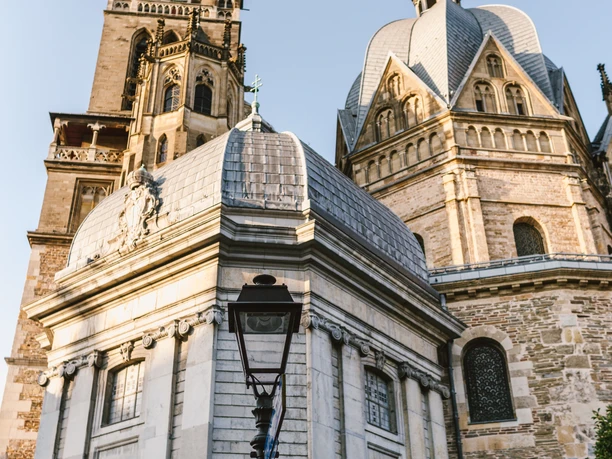 Aachener Dom vom Münsterplatz Der Aachener Dom erhebt sich majestätisch gegen den blauen Himmel, sichtbar sind der oktogonale Bau und die hohen Türme. Im Vordergrund bildet eine blühende Laterne einen farblichen Akzent.