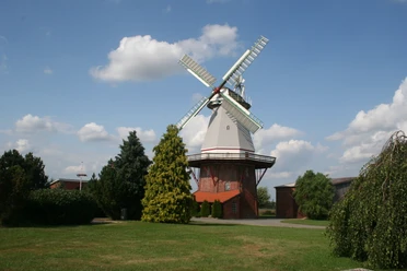 Windmühle unter blauem Himmel mit weißen Wolken, umgeben von grünen Bäumen und Wiesen.
