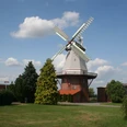 Blender Mühle Windmühle unter blauem Himmel mit weißen Wolken, umgeben von grünen Bäumen und Wiesen.