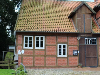 Fachwerkgebäude mit roten Ziegelwänden und Holztor, daneben drei weiße Fenster. Links ein Holzbalken.