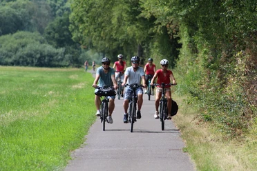 Two men and a woman are cycling along a paved path through a green landscape.