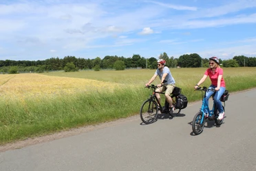 Two cyclists ride on a paved road through a green landscape in sunny weather.