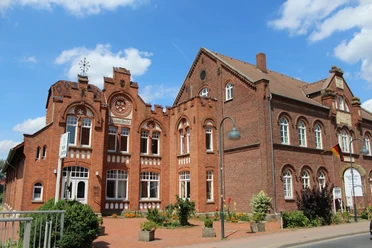 Historisches Backsteingebäude mit Spitzfenstern und Turmspitze, flankiert von Blumendekorationen.