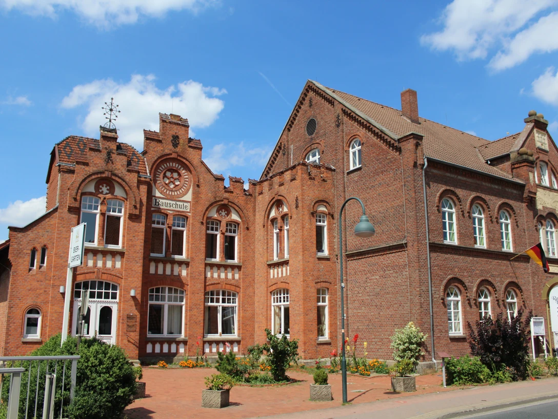 Bauschule Rehburg Historisches Backsteingebäude mit Spitzfenstern und Turmspitze, flankiert von Blumendekorationen.