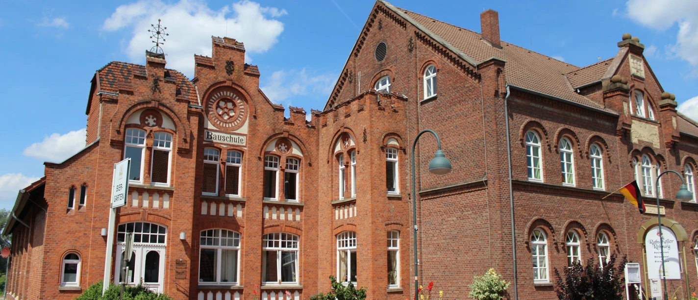 Bauschule Rehburg Historisches Backsteingebäude mit Spitzfenstern und Turmspitze, flankiert von Blumendekorationen.