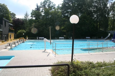 Wiehe-Bad Außenansicht eines großen Freibads mit breitem Wasserbecken, umgeben von grünen Bäumen und Sonnenterrassen.