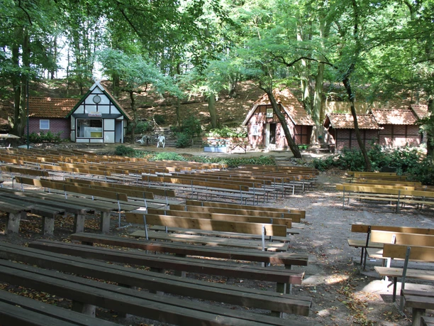 An open-air stage with wooden benches, surrounded by trees and half-timbered buildings in a rural setting.