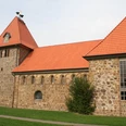 Historische Backsteinkirche mit roten Ziegeldächern und markantem Kirchturm, eingebettet in grüne Wiese.