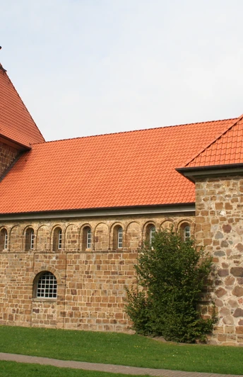 Historische Backsteinkirche mit roten Ziegeldächern und markantem Kirchturm, eingebettet in grüne Wiese.