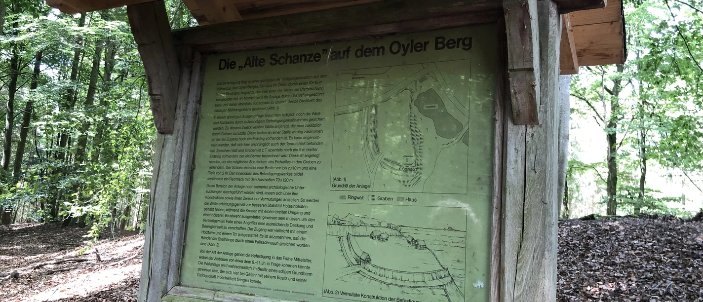 Information board in the forest with historical map and text about the "Alte Schanze" on Oyler Berg in the background.