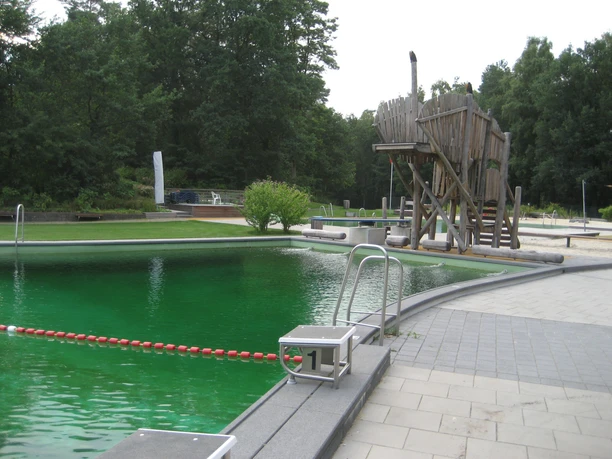 NaturWaldBad Steimbke Das NaturWaldBad Steimbke mit grünem Wasser und einem Holzspielschiff, umgeben von Natur.