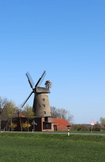 Historische Pottmühle Petershagen mit Windmühlenflügeln, umgeben von roten Gebäuden und grüner Landschaft.