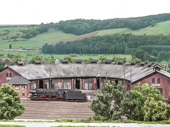 Modellbundesbahn Lokschuppen Ottbergen Historischer Lokschuppen Ottbergen mit Dampflok, umgeben von sanften grünen Hügeln und Bäumen.